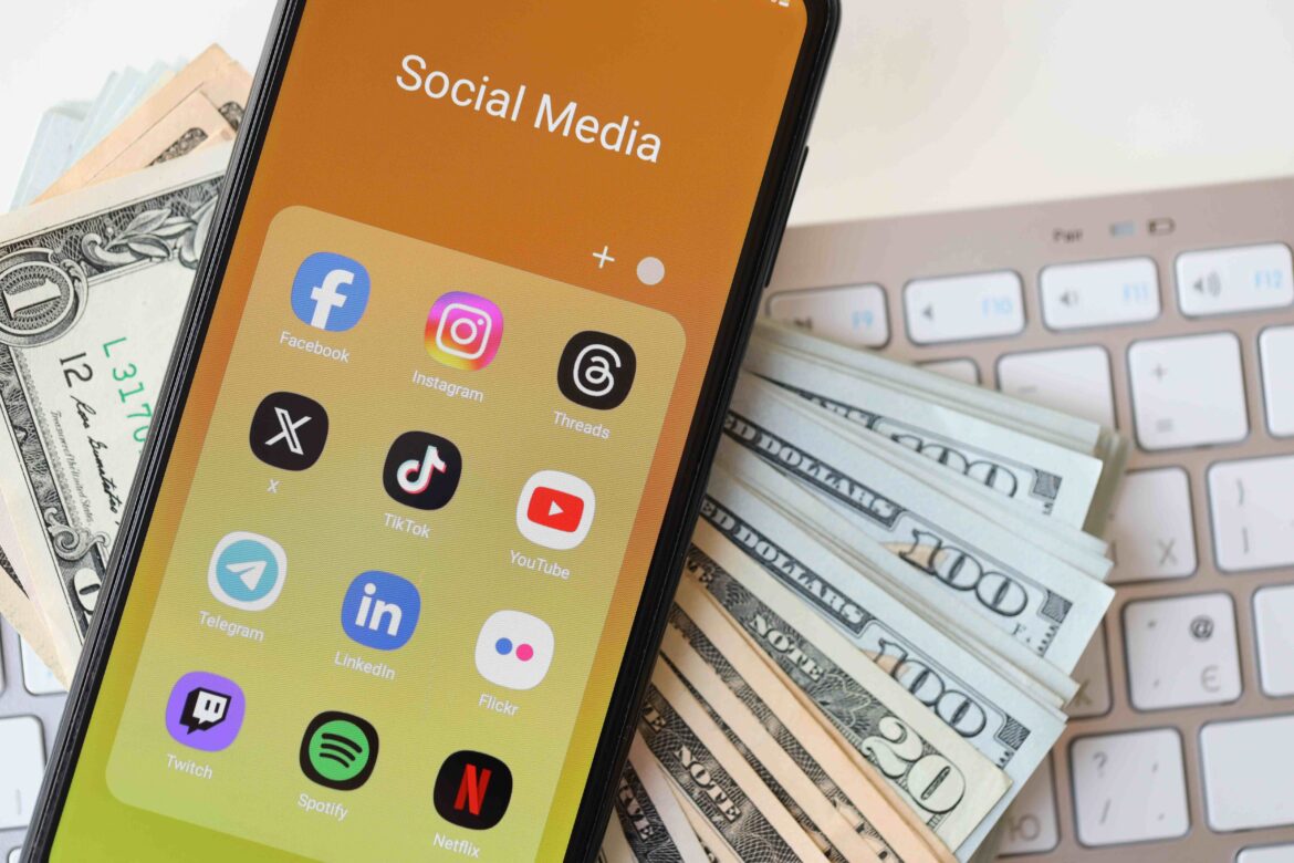 Many social media app icons on a smartphone screen with keyboard and cash in the background representing Social media influencing financial decision