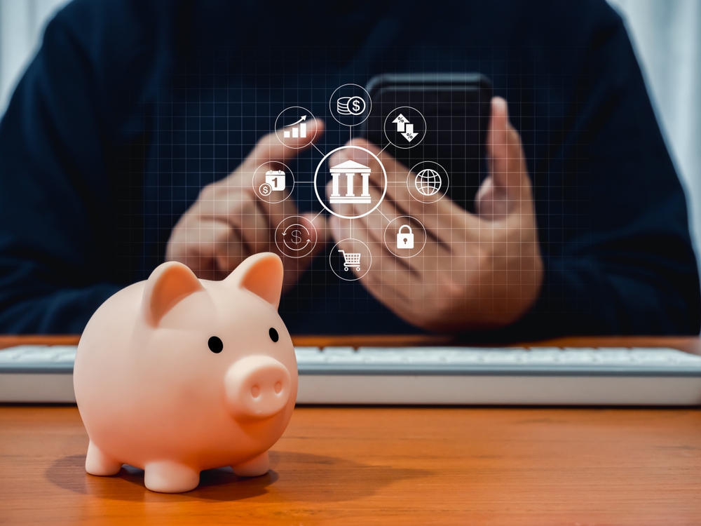 Online banking concept, saving account, financial transaction on mobile phone. Banking transaction data icons appear while person man using smartphone application with pink piggy bank on wooden table.
