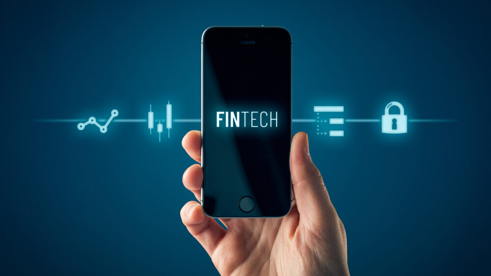 Fintech written on mobile screen held by a hand and virtual representation of finance technology in the background